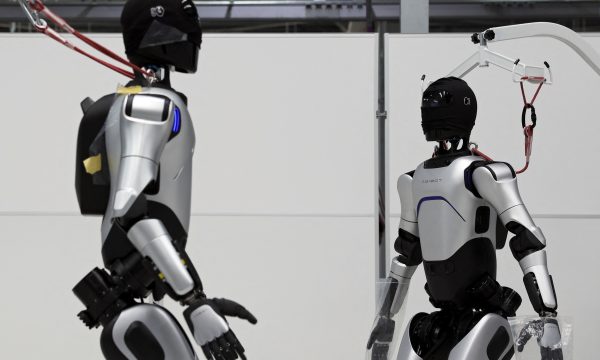 Dos robots humanoides plateados están siendo probados en un laboratorio industrial, conectados a un sistema de suspensión.