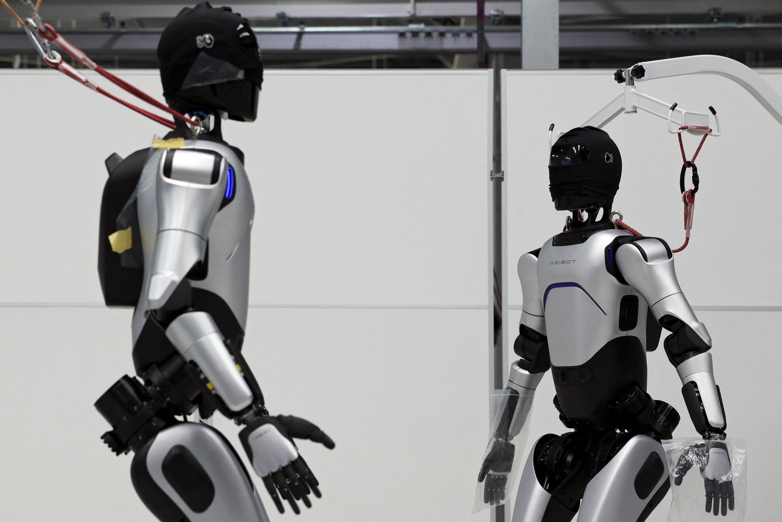 Dos robots humanoides plateados están siendo probados en un laboratorio industrial, conectados a un sistema de suspensión.