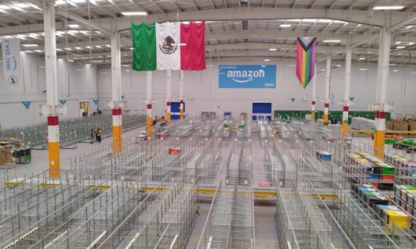 Interior del nuevo centro logístico DXX2 de Amazon en la alcaldía Coyoacán, Ciudad de México, con estanterías metálicas vacías y banderas institucionales.