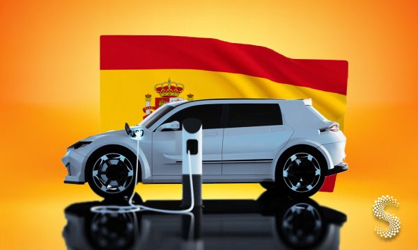 Coche eléctrico cargando frente a la bandera de España, símbolo del crecimiento de matriculaciones de vehículos híbridos y eléctricos en 2025.